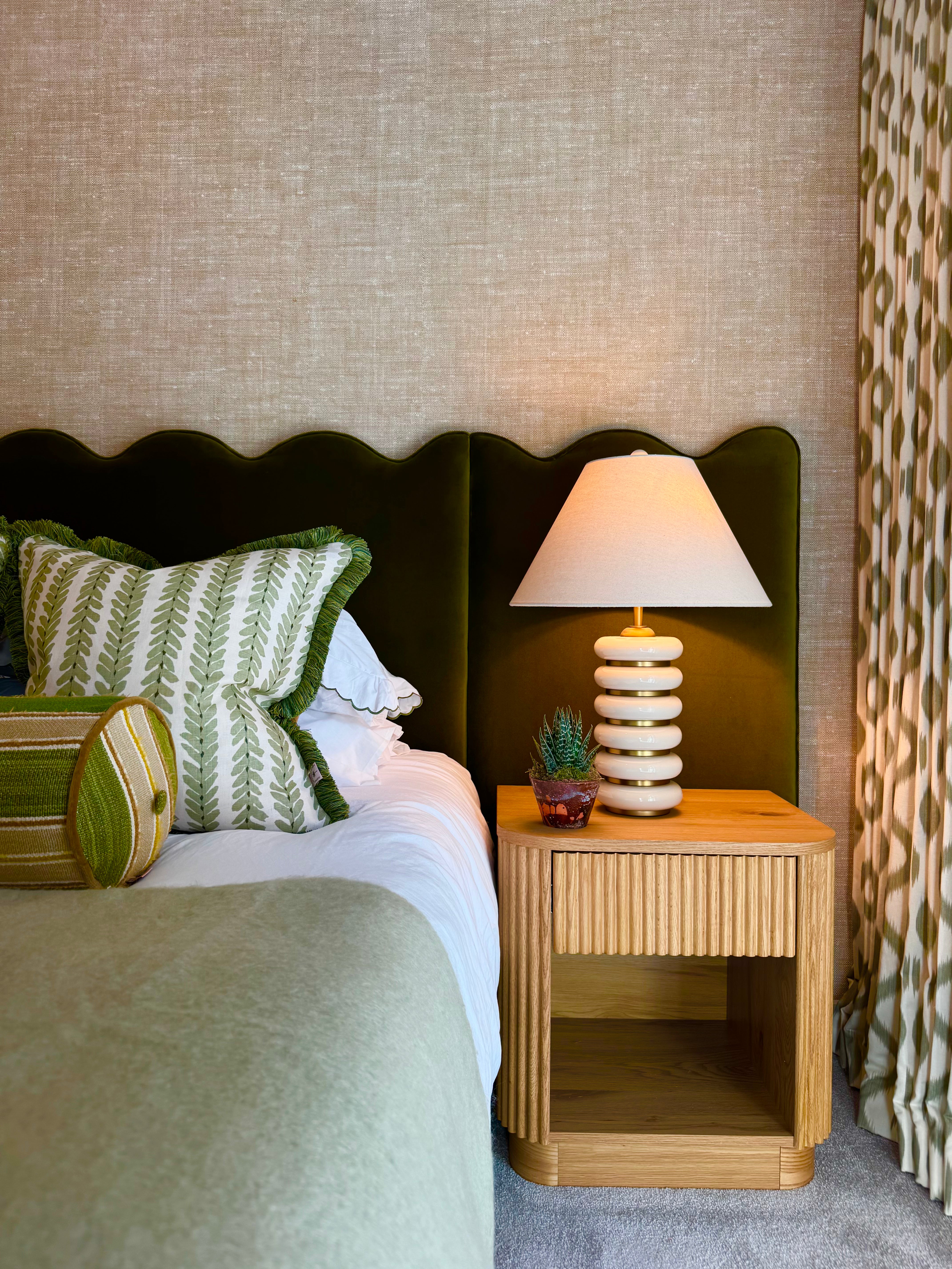 Bedroom with green upholstered headboard, patterned pillows, and wooden nightstand with lamp.